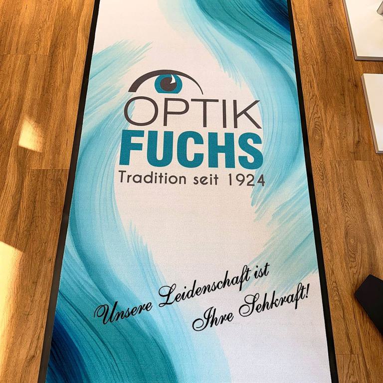 Werbematte von Optik Fuchs mit blauem Design auf Holzboden.