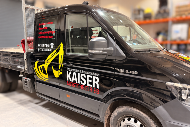 Schwarzer Firmenwagen mit Pritsche in einer Halle, beschriftet mit Kaiser Meisterbetrieb und Baggergrafik.