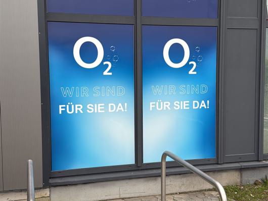 Schaufenster mit großer O2-Werbung an einem Gebäude neben Gehweg und Absperrband