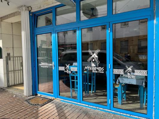Blaue Glasfront eines Restaurants mit dem Schriftzug Mykonos Greek Food und Tischen im Inneren.