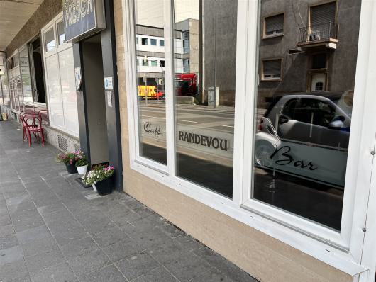 Straßenansicht eines Cafés mit großen Fenstern, Blumentöpfen und roten Stühlen auf dem Gehweg