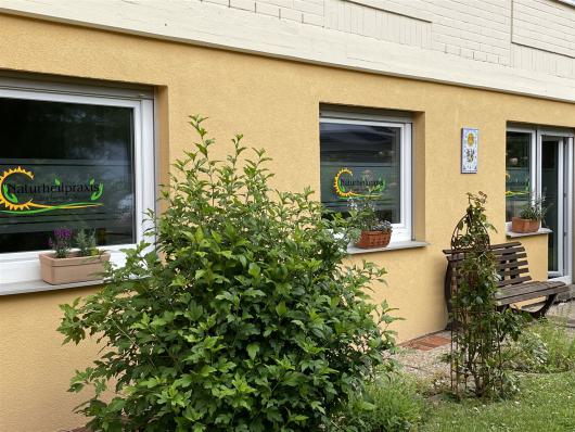 Eingang einer Naturheilpraxis an einem gelben Haus mit Fenstern, Bank und Sträuchern davor