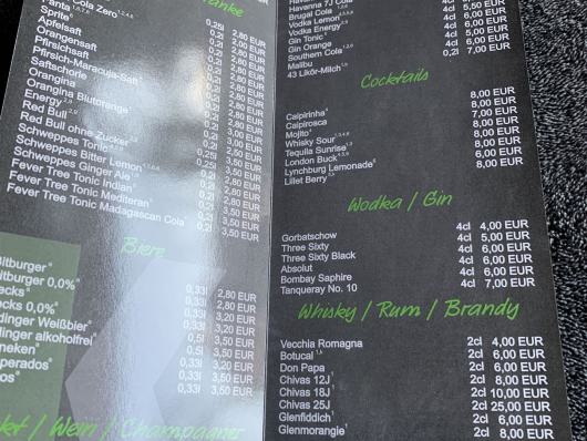 Aufgeklappte Getränkekarte auf dunklem Stoff mit Preisen für Softdrinks, Bier, Cocktails und Shots.