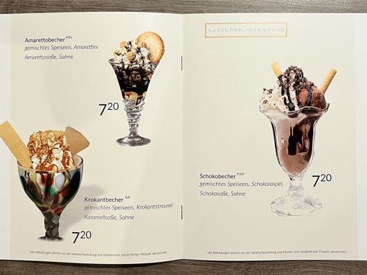 Aufgeschlagene Eiskarte mit Fotos von Eisbechern und Preisen auf einer Holzfläche.