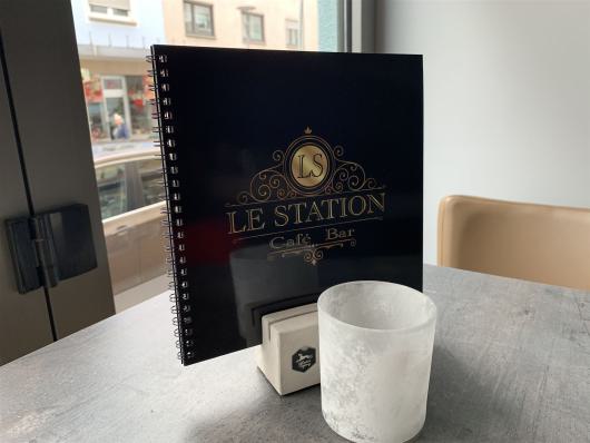 Schwarze Speisekarte von Le Station auf einem Tisch am Fenster neben einem weißen Glas