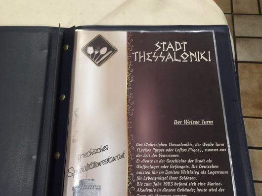 Aufgeschlagene Speisekarte mit einer Seite über Stadt Thessaloniki und den Weißen Turm auf einem Tisch.