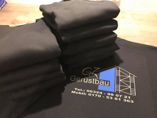 Mehrere gefaltete schwarze Textilien mit weißem Gerüstbau-Aufdruck liegen gestapelt auf einem Tisch.