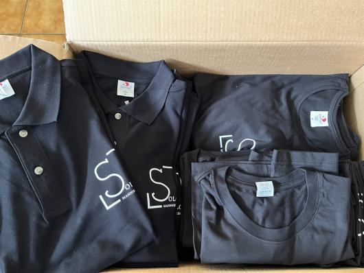 Geöffneter Karton mit gefalteten dunklen Poloshirts und T-Shirts mit weißem Logo.