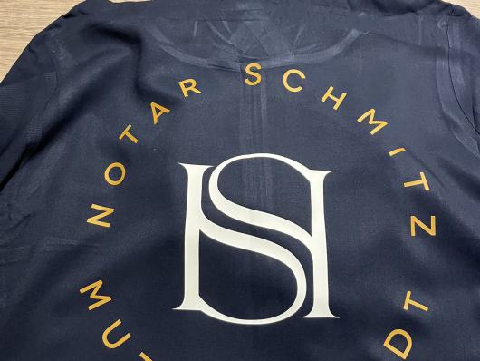 Dunkles T-Shirt mit großem Logo und kreisförmigem Schriftzug auf Holzboden.