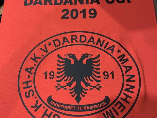 Rotes Kleidungsstück mit dem schwarzen Aufdruck Dardania Cup 2019 und einem runden Vereinslogo