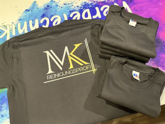 Mehrere dunkelgraue T-Shirts mit dem Aufdruck MK Reinigungsprofi liegen auf einer bunten Fläche.