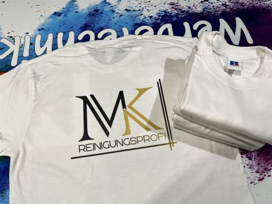Weißes T-Shirt mit dem Aufdruck MK Reinigungsprofi neben einem Stapel weiterer Shirts.