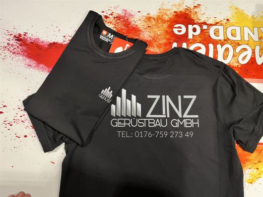 Zwei schwarze T-Shirts mit silbernem Firmenaufdruck liegen auf einer farbig bespritzten Fläche.