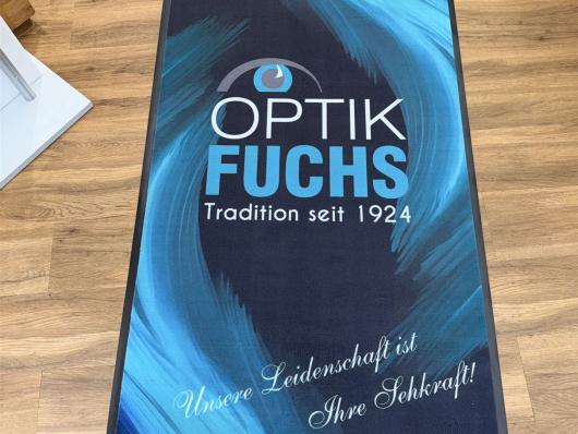 Dunkle Bodenmatte mit blauem Muster und dem Schriftzug Optik Fuchs auf Holzfußboden
