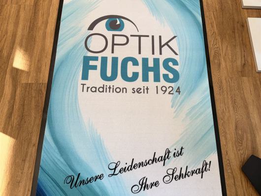 Große Werbematte von Optik Fuchs auf einem Holzboden