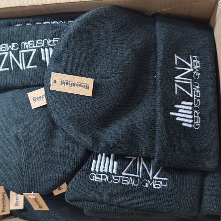 Mehrere schwarze Strickmützen mit aufgesticktem Firmenlogo in einem Karton.