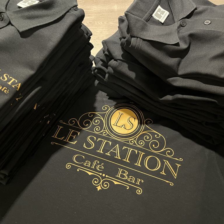 Mehrere gefaltete schwarze Poloshirts mit goldenem Logo auf einem Tisch.