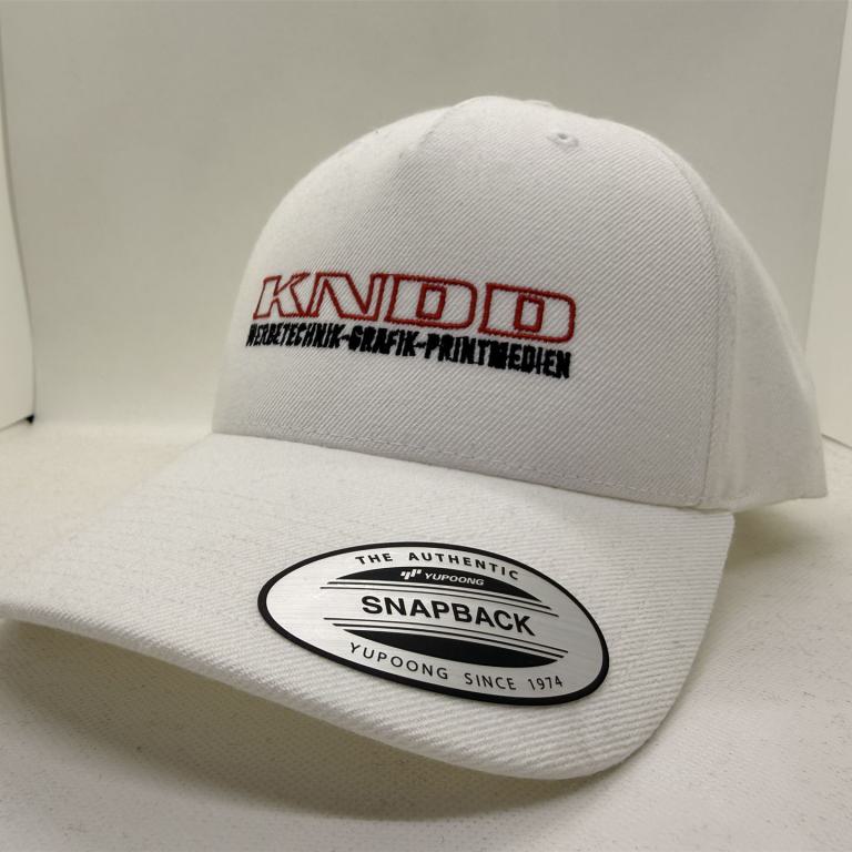 Weiße Snapback-Kappe mit aufgesticktem KANDO-Logo und Aufkleber auf dem Schirm.
