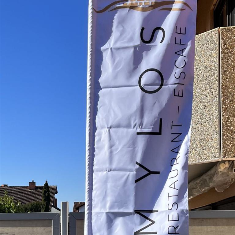 Weiße Werbefahne mit der Aufschrift MYLOS Restaurant Eiscafe an einem Gebäude vor blauem Himmel.