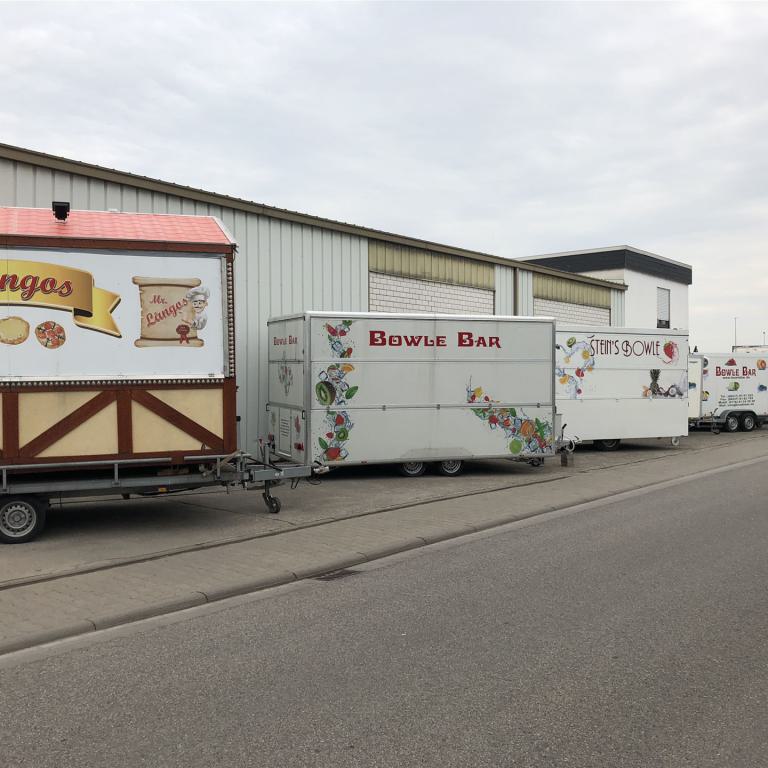 Mehrere Imbissanhänger stehen entlang einer Straße vor einer Lagerhalle.