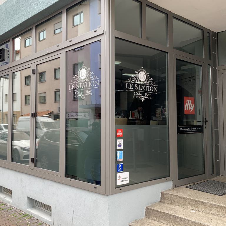 Eingangsbereich des Cafés Le Station mit großen Glasfenstern an einer Straßenecke