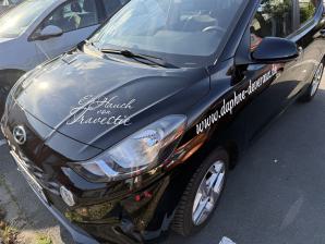 Schwarzes Auto mit Werbeschrift auf Motorhaube und Seite auf einem Parkplatz